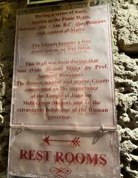 Stone wall with two white signs with red writing. The top one gives a brief history of Roman Malta and then states “The Roman senator and orator Cicero commented on the importance of the Temple of Juno on Melita (now Mdina), and on the extravagant behaviour of the Roman governor.” The second sign points to the restroom.